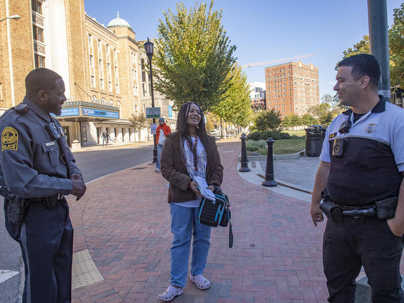 A woman standing between to VCU Police Officers on a sidewalk next to a park. 