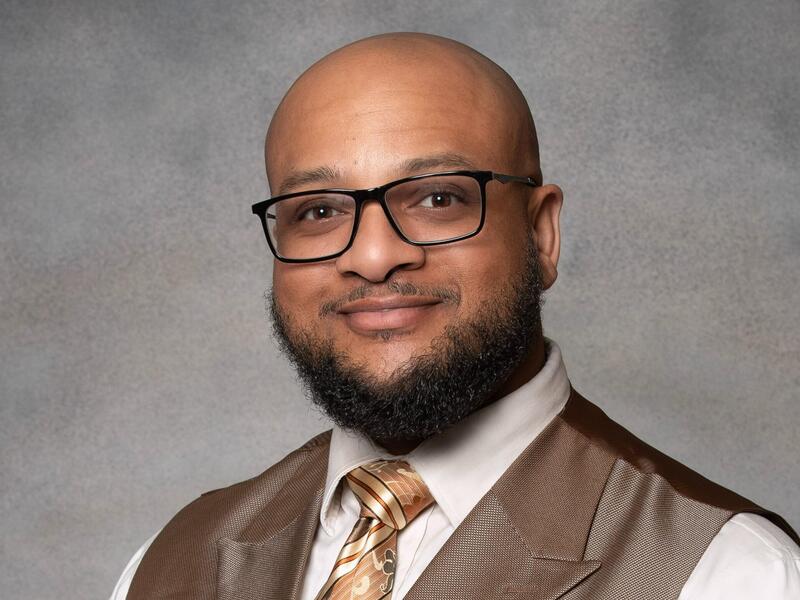 A photo of a man from the chest up. He is wearing a vest, button up shirt, tie, and glasses. 