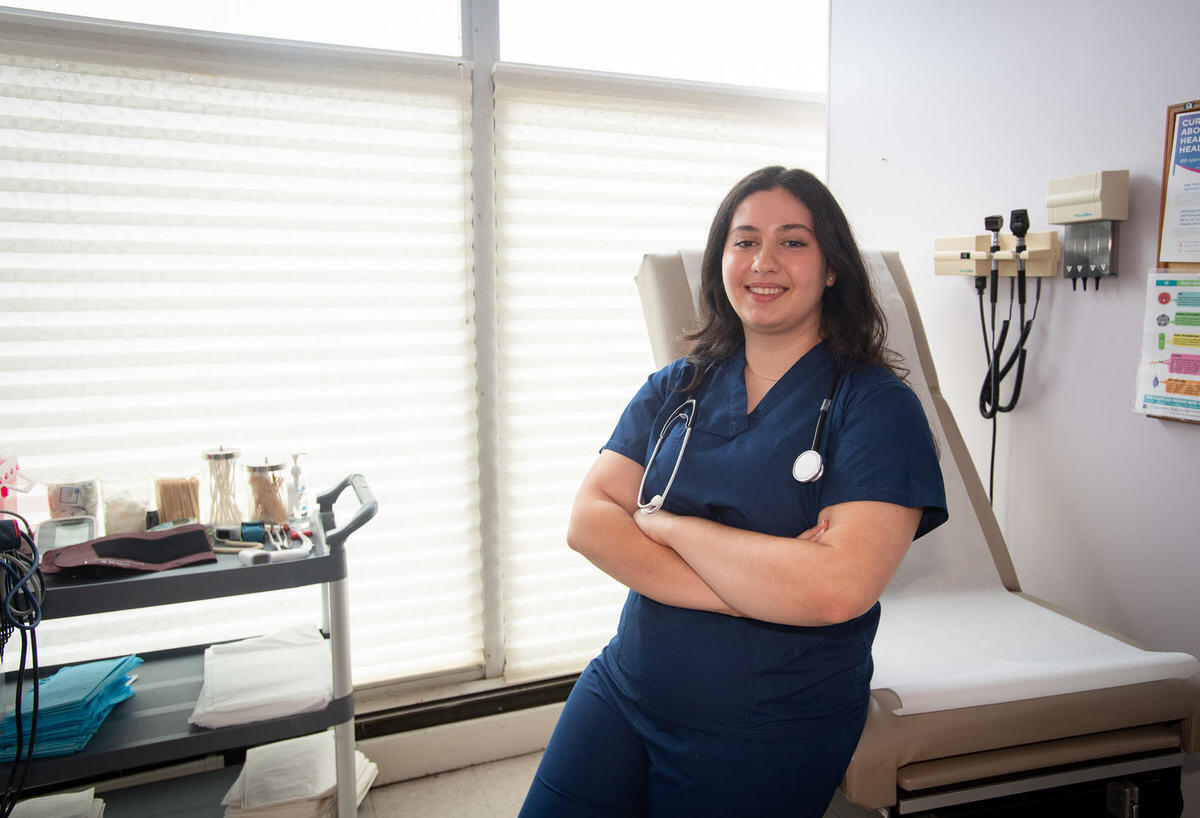 A photo of a woman wearing medical scrubs from the waist up. She is leaning against an examination table with her arms cross against her chest. 