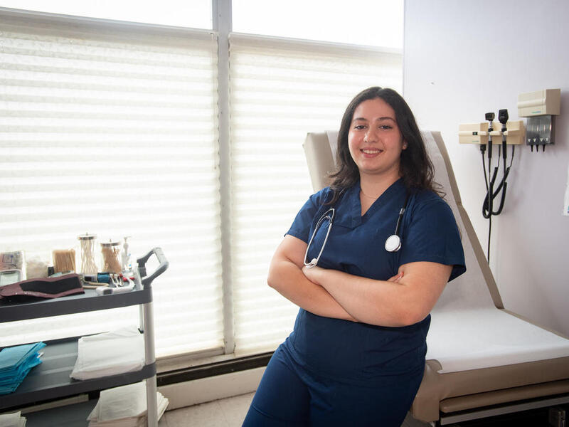 A photo of a woman wearing medical scrubs from the waist up. She is leaning against an examination table with her arms cross against her chest. 
