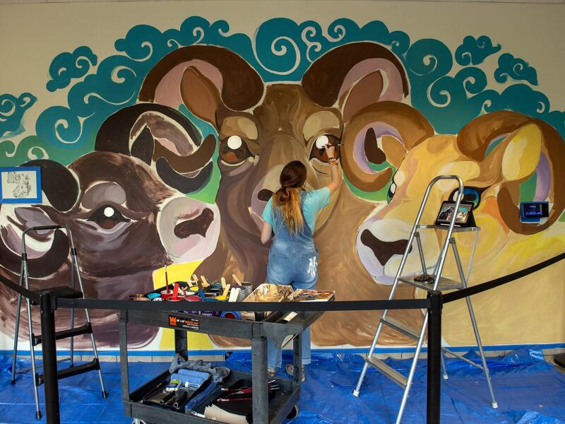 A photo of a woman paiting a mural with of three rams. 