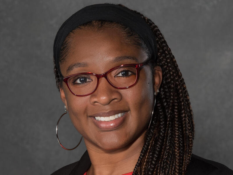 A portrait of a woman with long braids wearing a black cardigan, glasses, and hoop earrings. 
