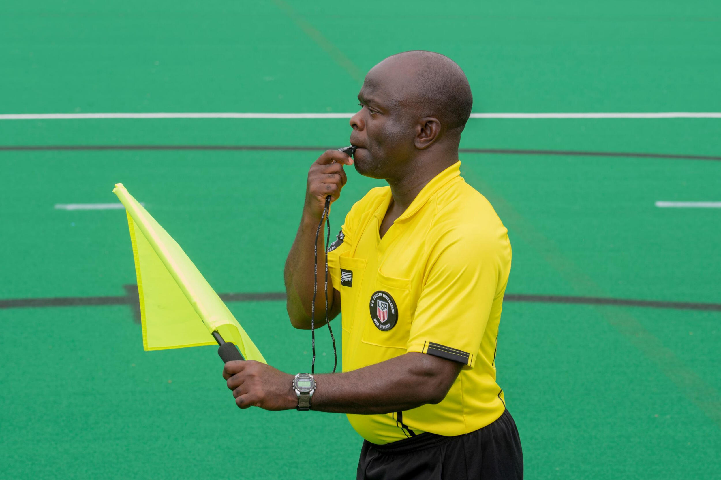 Kwasi Deitutu in soccer ref uniform holds a flag and blows a whistle.