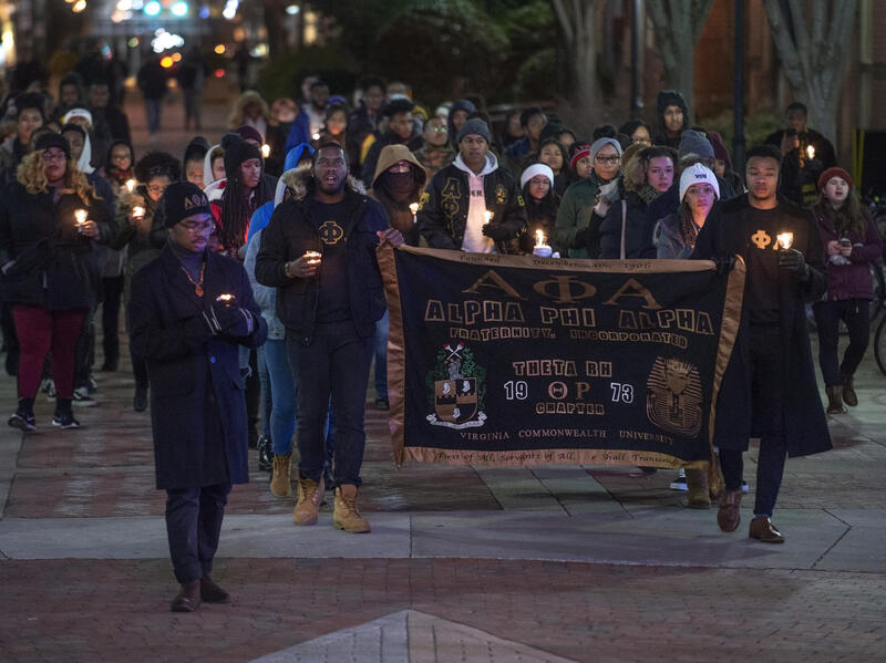 A photo of a group of people walking with candles. Two people in the front of the group hold up a banner for Alpha Phi Alpha. 