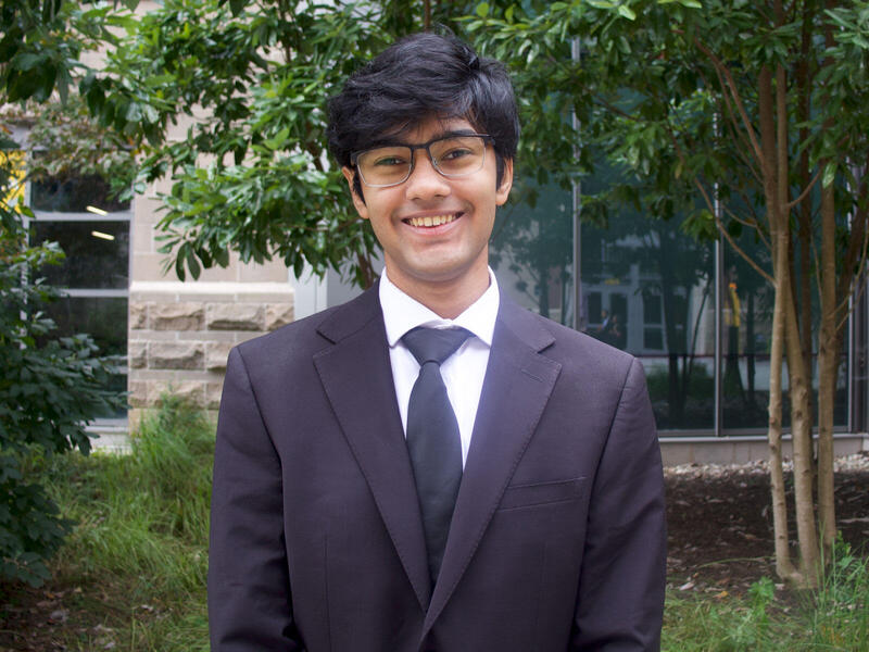A photo of a man from the chest up. He is wering a black suit and tie, and a white button down shirt. 