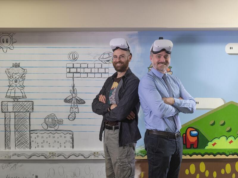 Two smiling men standing back to back with their arms crossed wearing virtual reality goggles headsets on their foreheads 