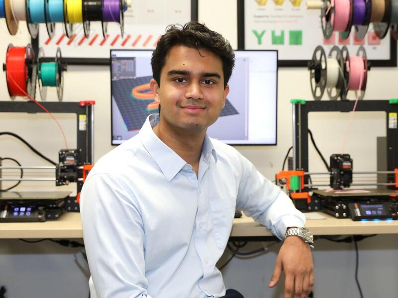 A photo of a man sitting at a desk with a computer screen and two 3D printers on it. 