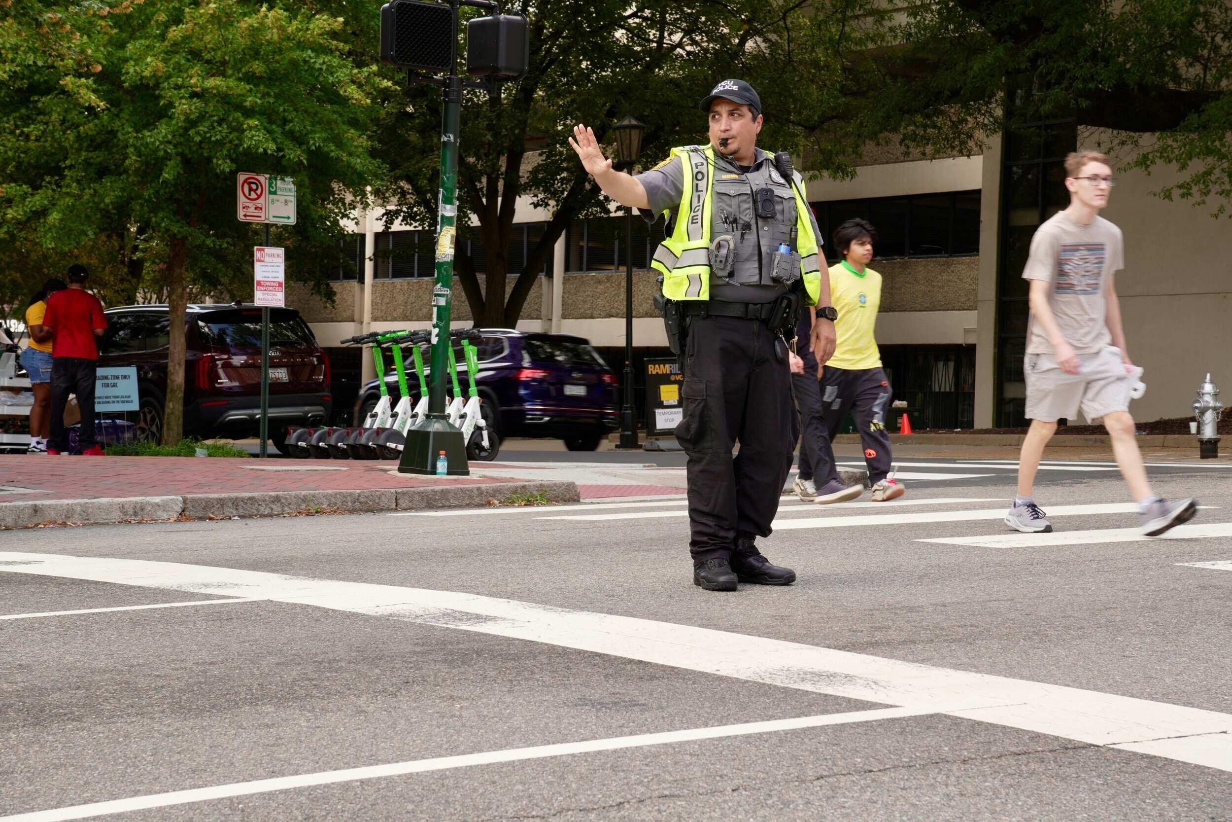 A photo of a police officer standing at a crosswalk and holding his left hand up in a \"stop\" motion. 