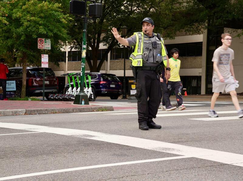 A photo of a police officer standing at a crosswalk and holding his left hand up in a \"stop\" motion. 