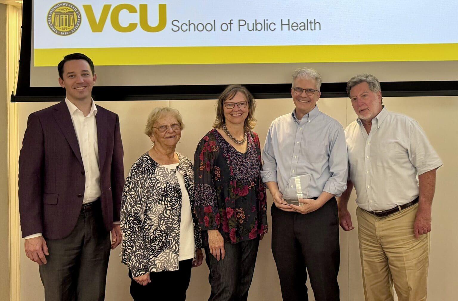 A group photo of five people standing. Behind them is a screen that says \"VCU\" in yellow letters and \"School of Public Health\" in black letters. 