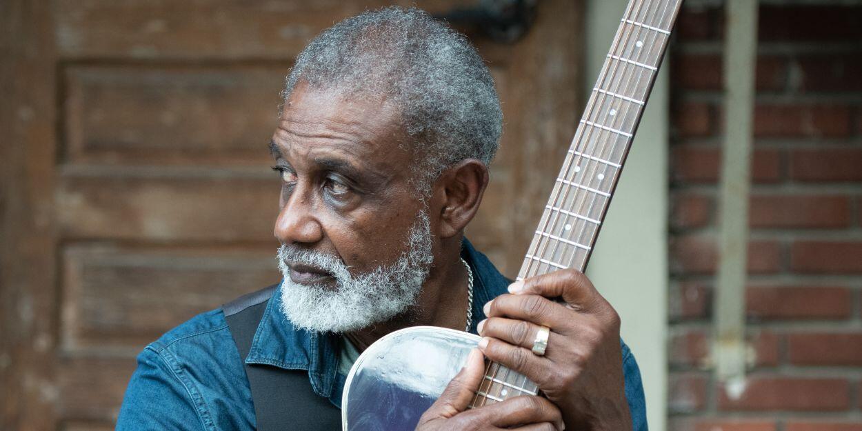 A man with gray hair and a blue button up shirt looking to the left while holding an acoustic guitar. 