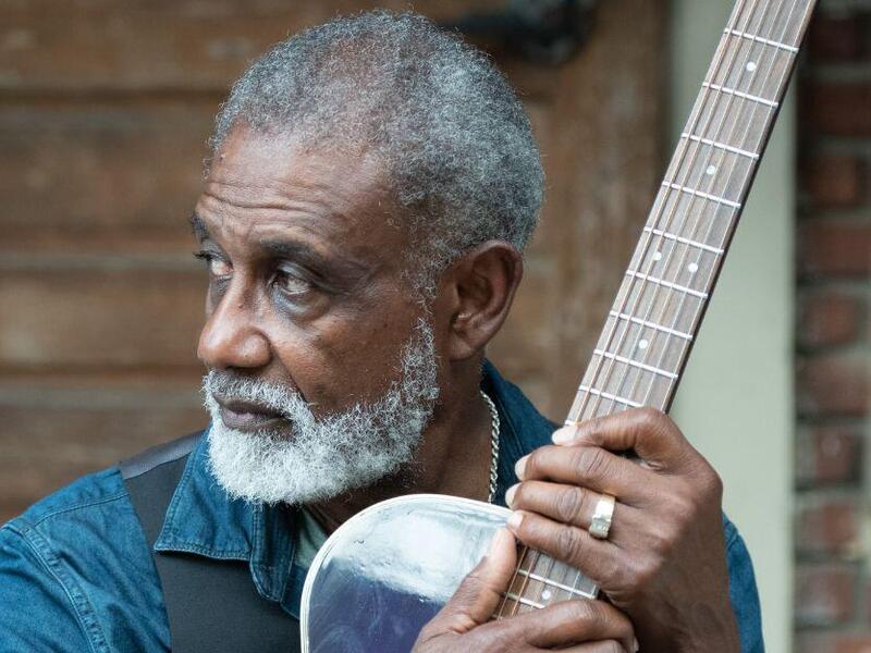 A man with gray hair and a blue button up shirt looking to the left while holding an acoustic guitar. 