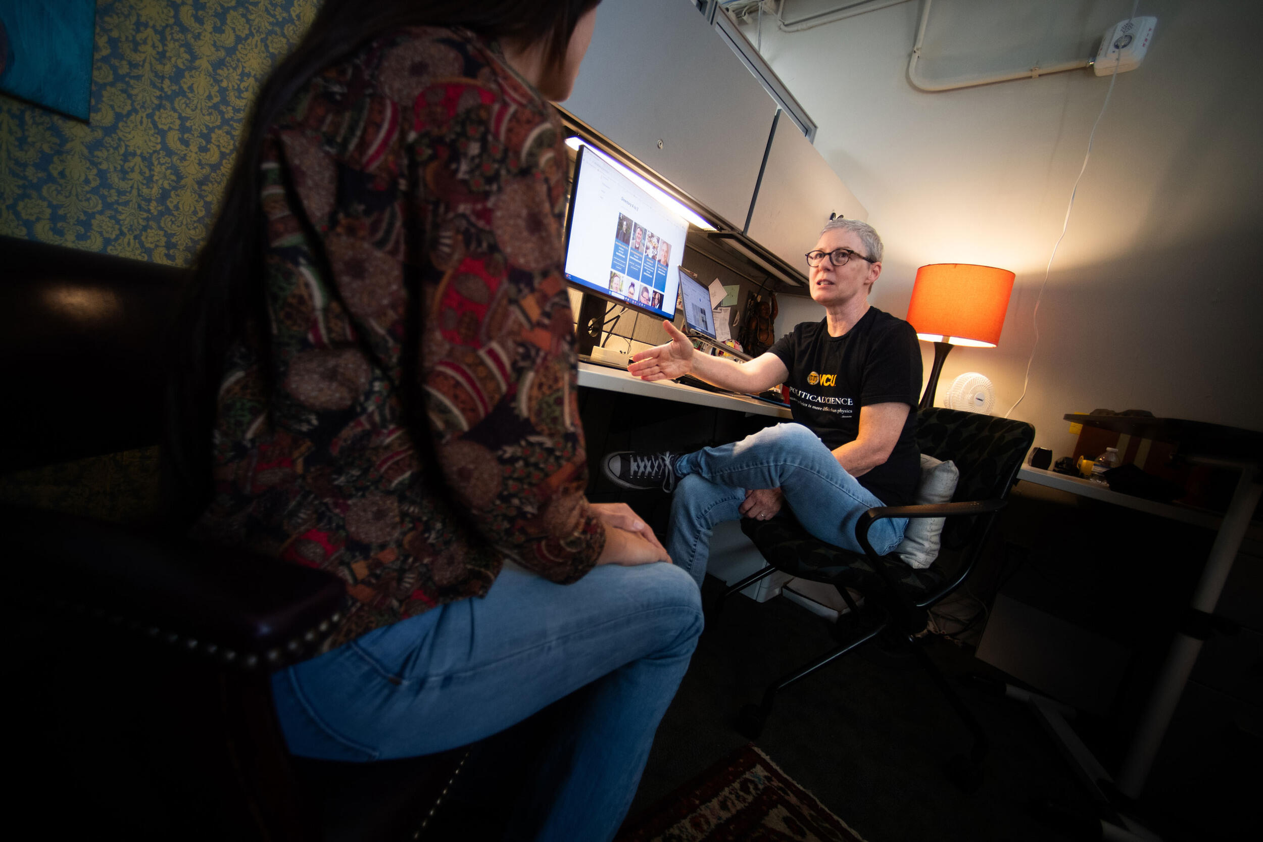A photo of a woman seated at a desk talking to a woman who is sitting in a chair across from it. 
