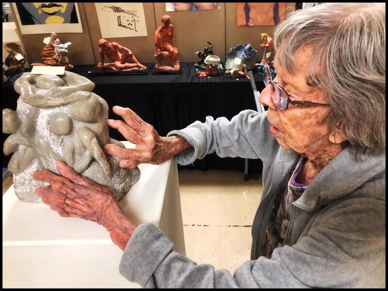 A photo of an old woman wearing glasses, touching a Sculpture. The sculpture is gray and shows several human figures moving around the side sides of it.