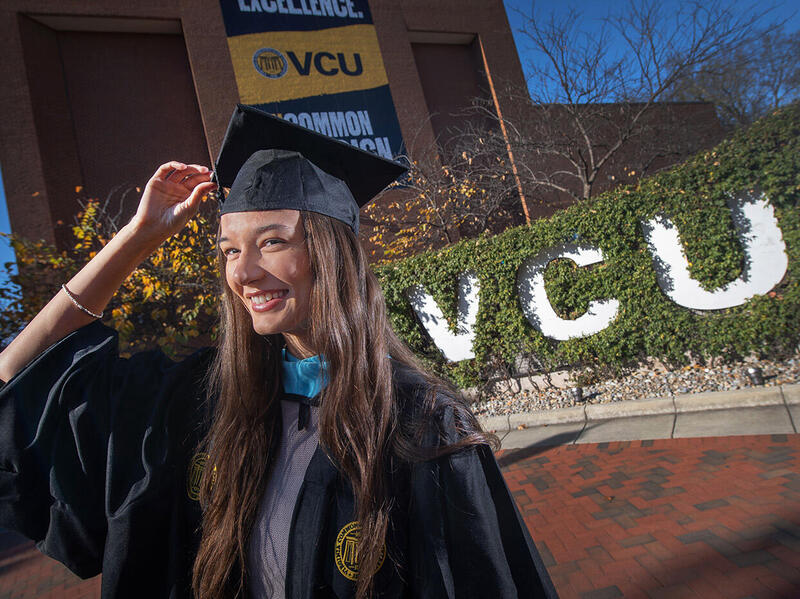 A photo of a woman wearing a graduation cap and gown standing in front of white letters that spell out \"VCU.\"