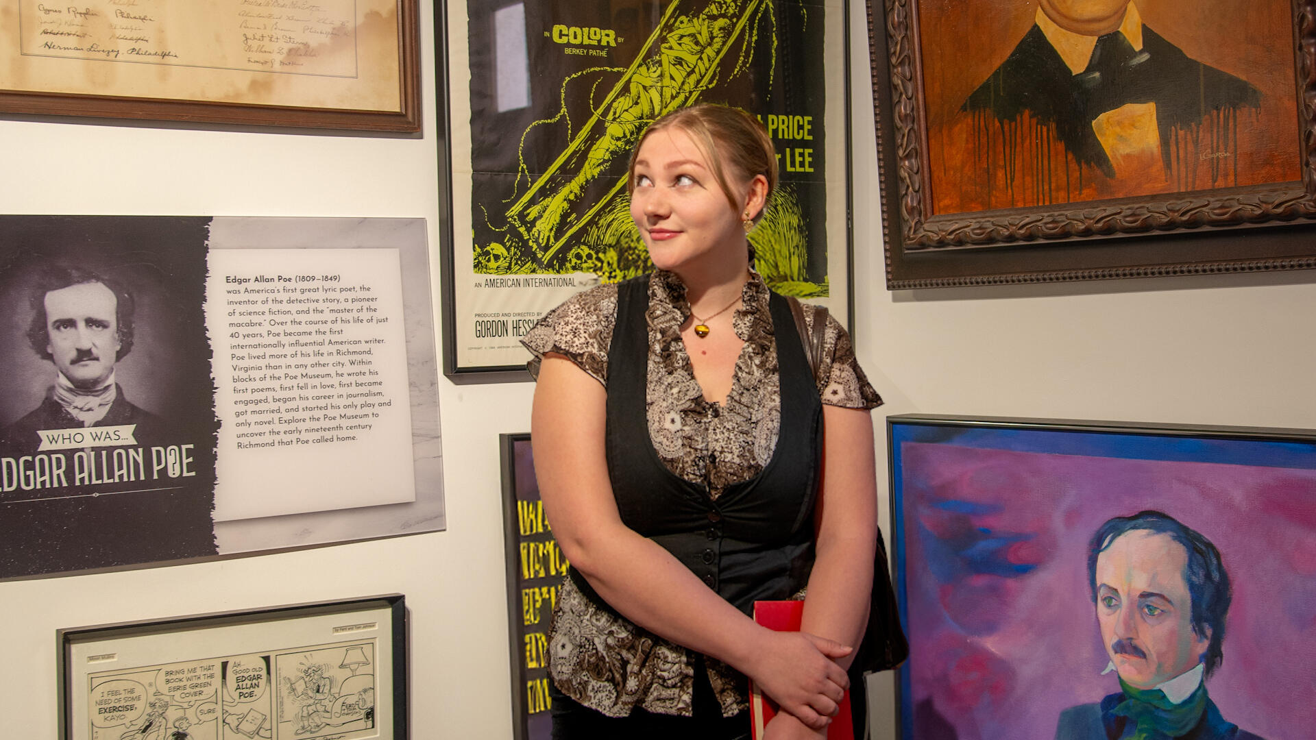 A photo of a woman from the waist up. She is standing in front of a gallery wall covered in illustrations of Edgar Allen Poe. 