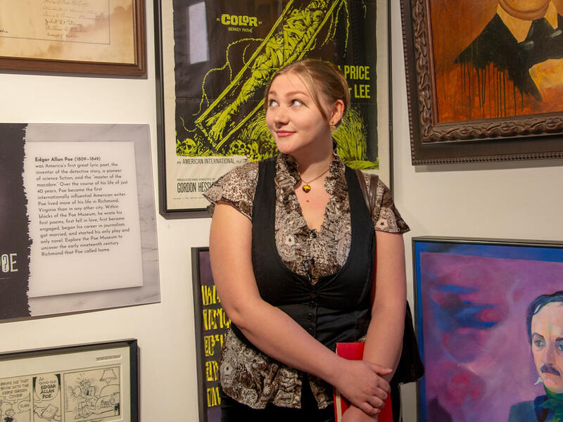 A photo of a woman from the waist up. She is standing in front of a gallery wall covered in illustrations of Edgar Allen Poe. 
