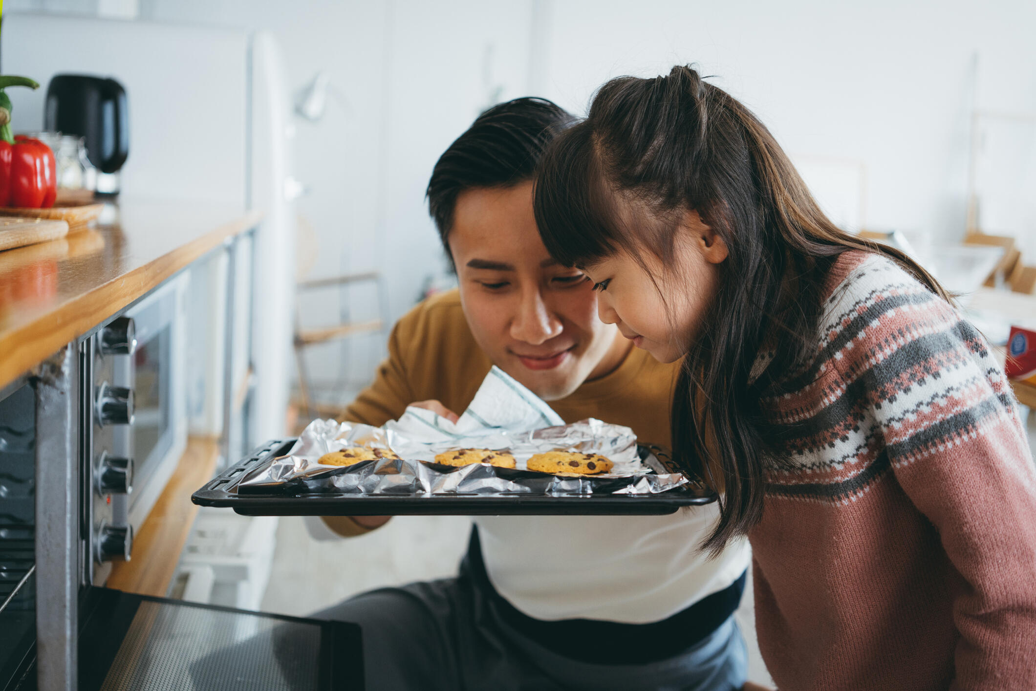 A man and a little girl smelling a tray of cookies in front of an oven. 