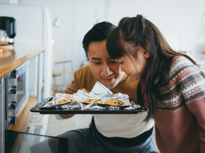 A man and a little girl smelling a tray of cookies in front of an oven. 