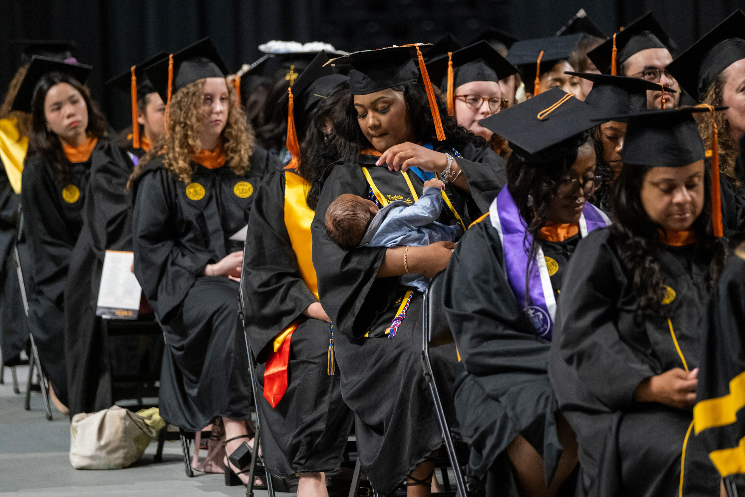 A photo of college graduates sitting in chairs wearing graduation caps and gowns. One woman on the edge of the third row is holding an infant. 