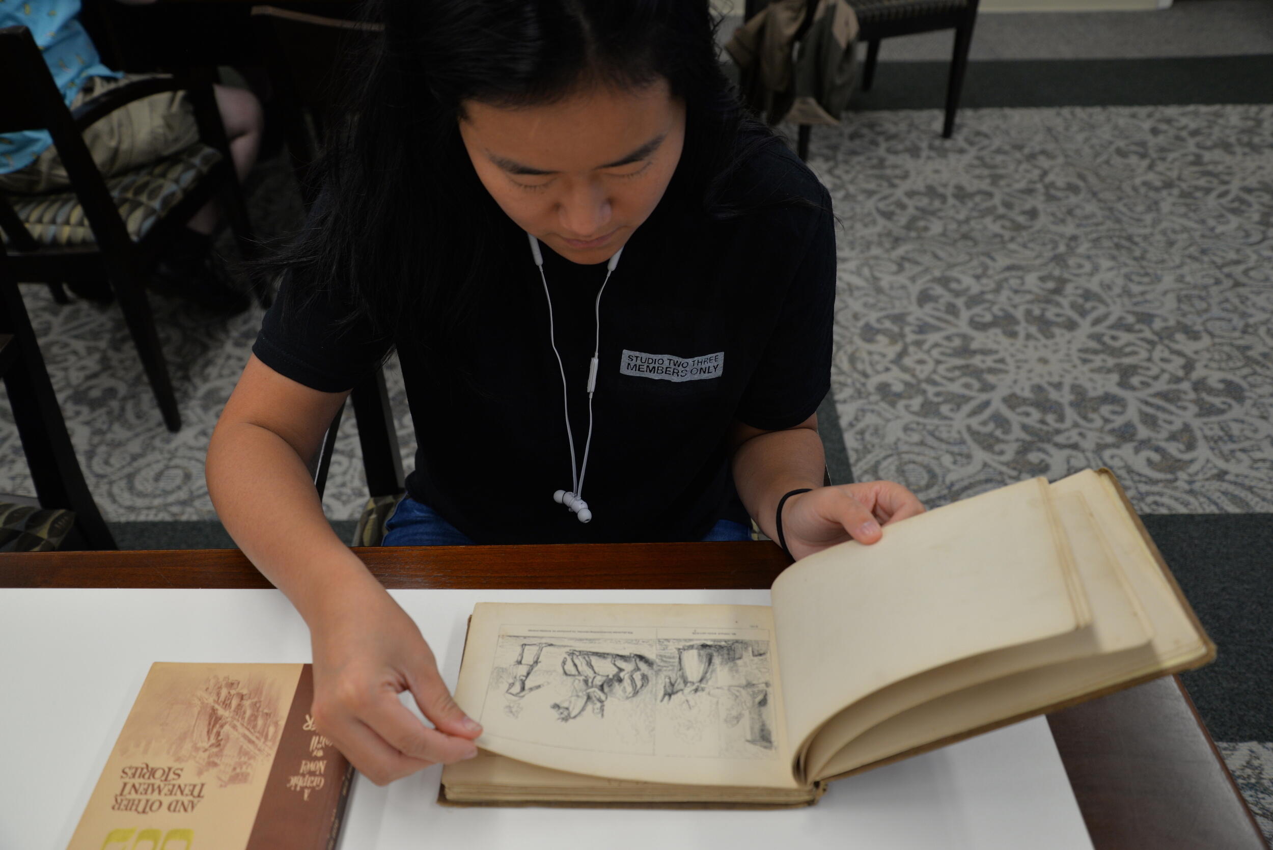 A student wearing headphones reading a book at a table.
