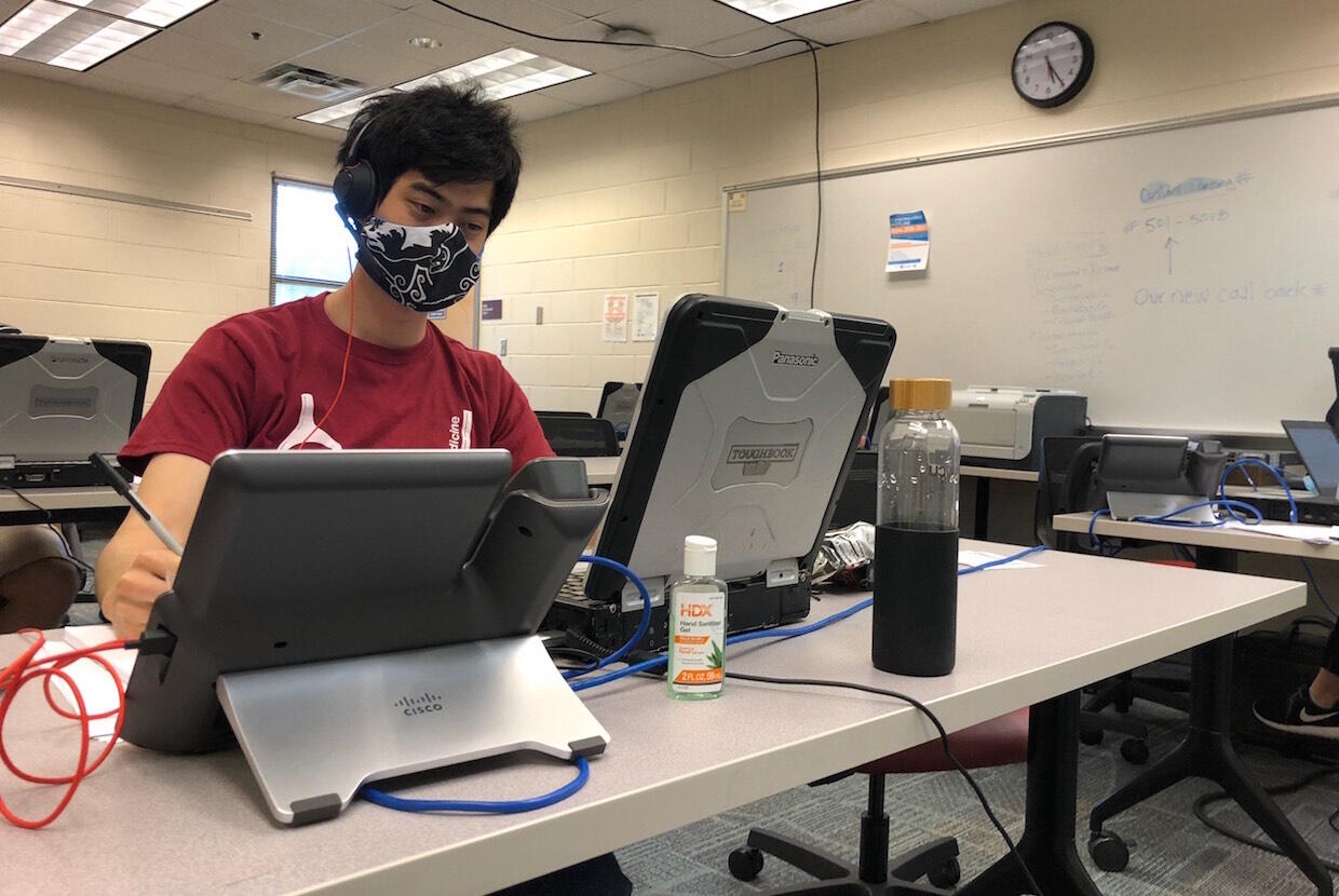 A person wearing a mask and seated at a classroom desk uses a laptop.