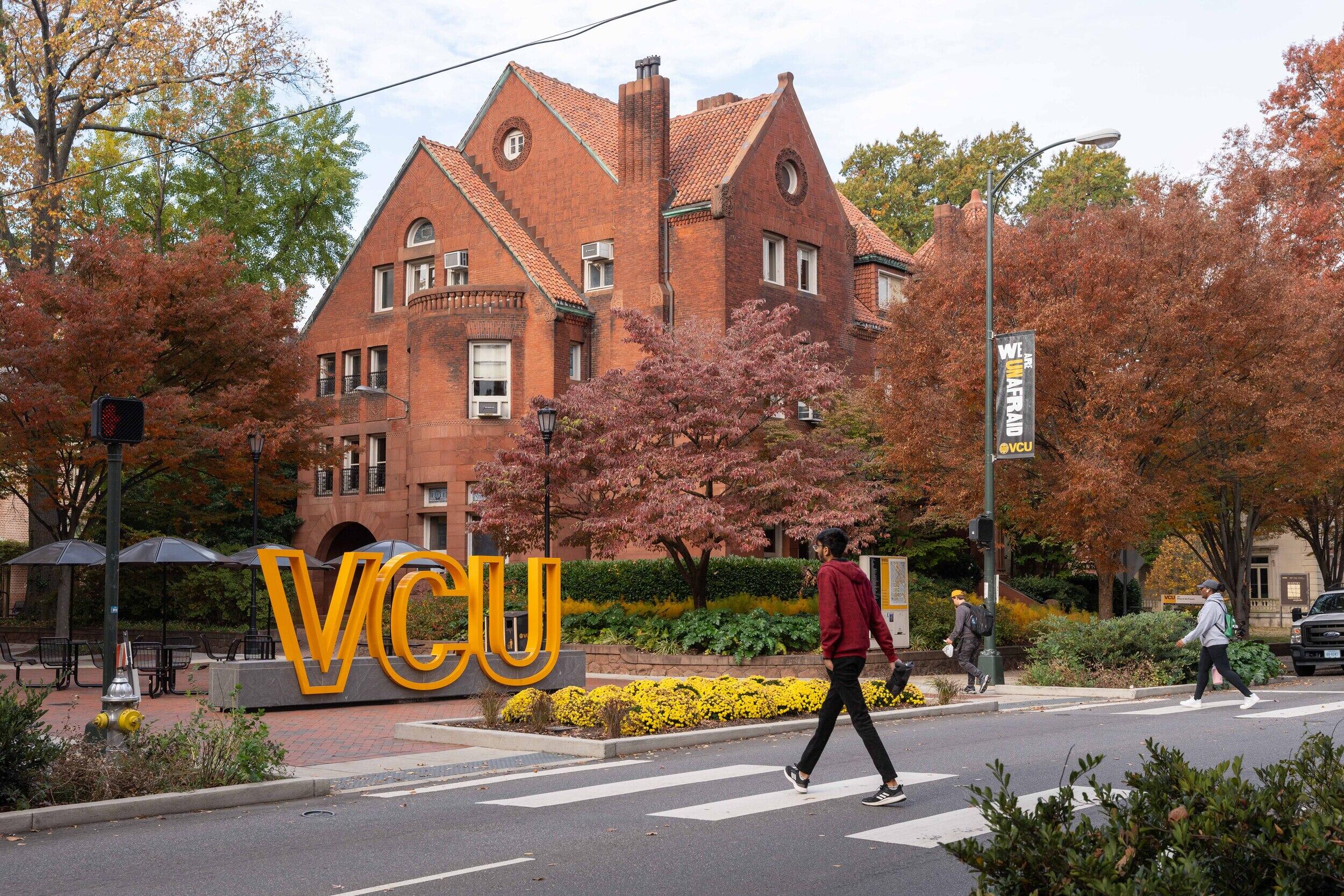 A photo of a person walking across a street with a cross walk. Onn the left side of the street is a large brick house and a sign that says \"VCU\" in large yellow letters. 