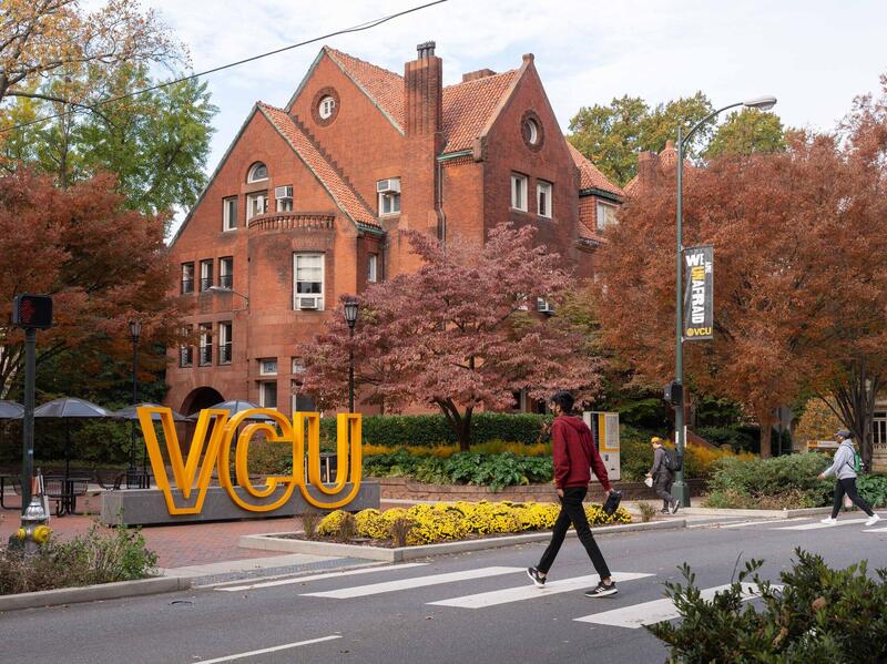 A photo of a person walking across a street with a cross walk. Onn the left side of the street is a large brick house and a sign that says \"VCU\" in large yellow letters. 