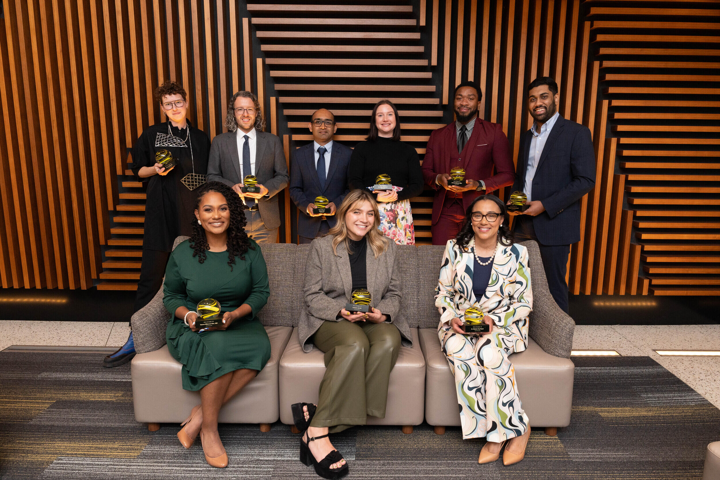 A group photo of nine people, three sitting on a couch and five standing behind the couch. 