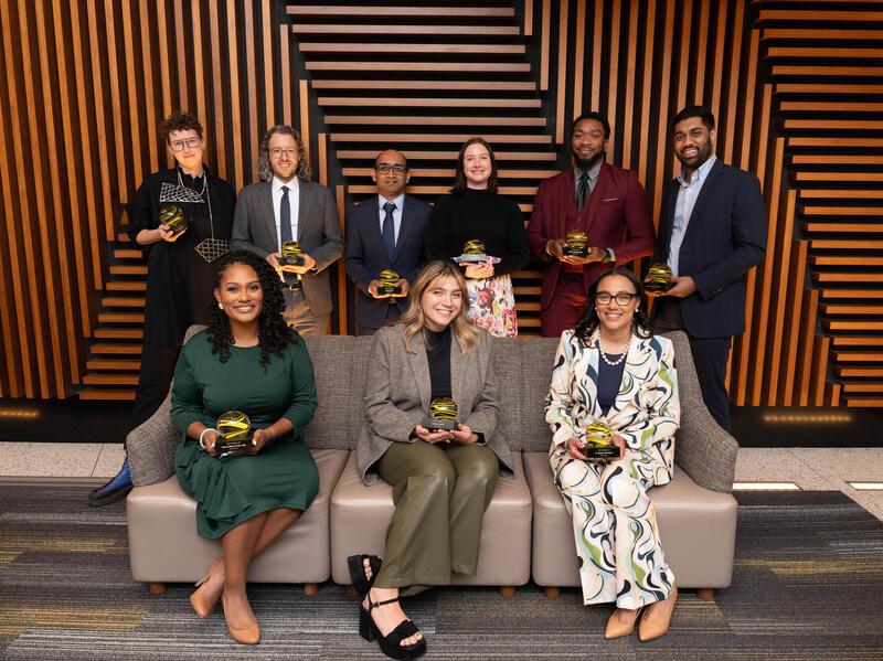 A group photo of nine people, three sitting on a couch and five standing behind the couch. 