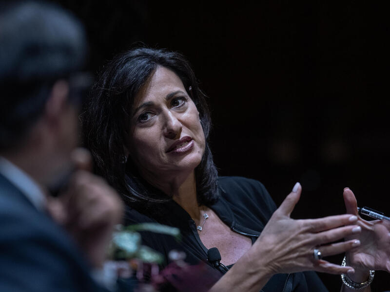 A photo of a woman speaking while gesturing her hands in front of her. She is looking at a man sitting to her left. 
