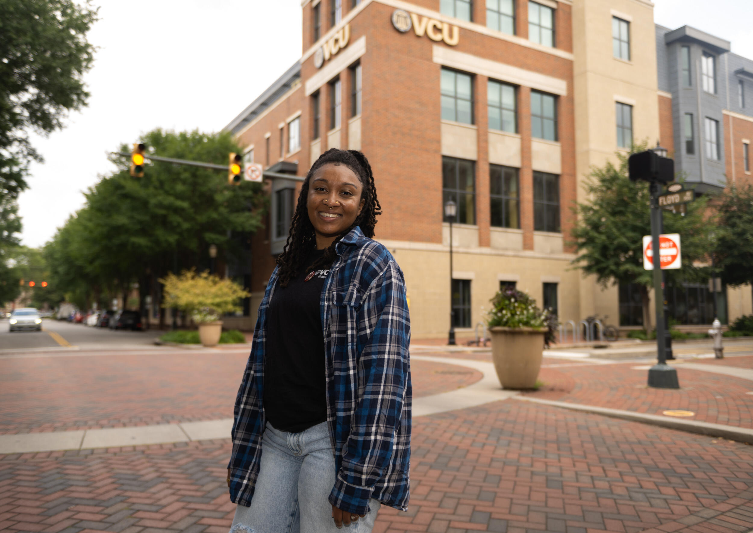 A woman in a flannel shirt smiles and stands in a courtyard with a building behind her.
