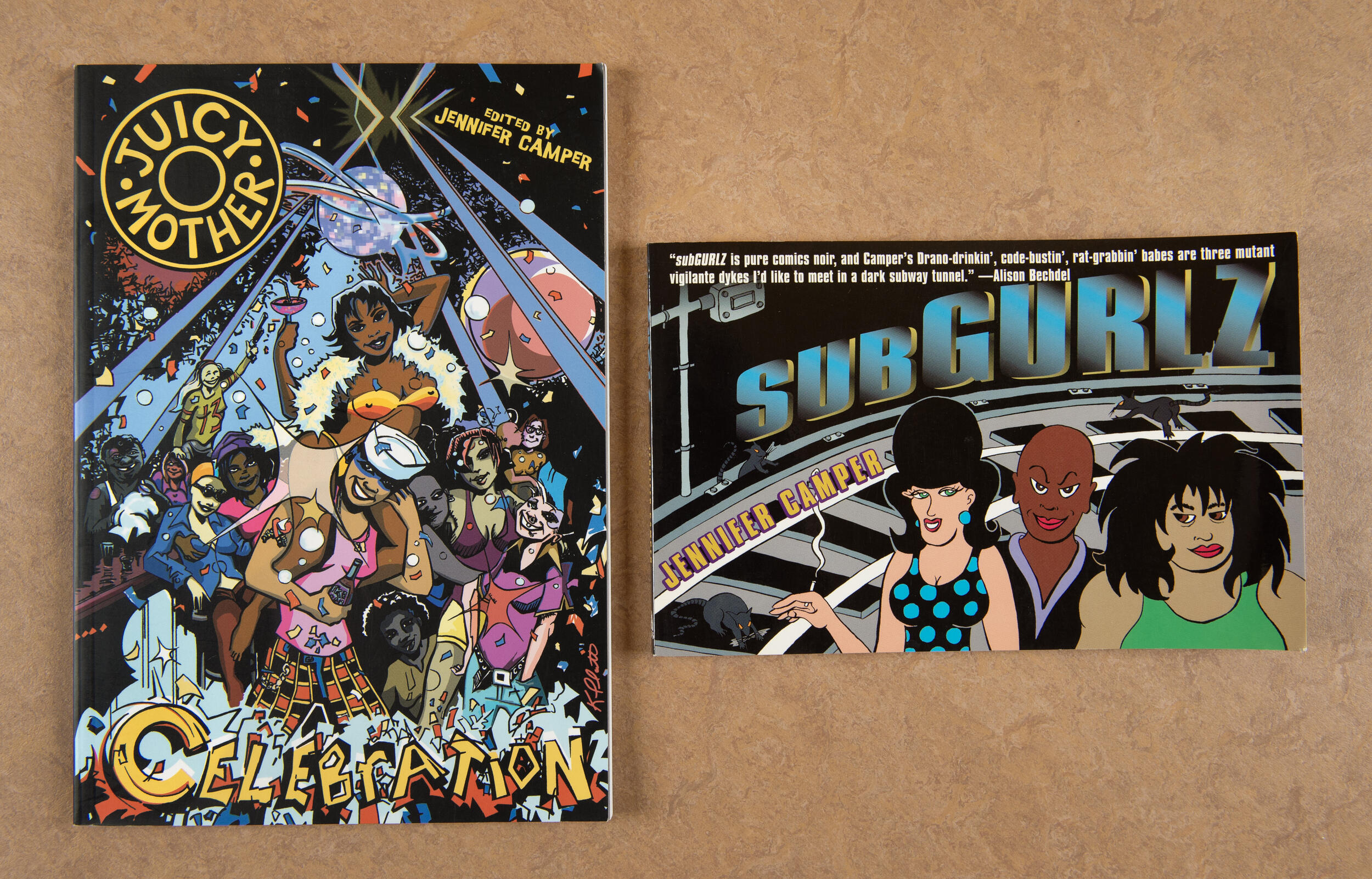 A photo of a comic book cover that has a group of people dancing at a gay club. In the upper left corner is a black circle with yellow text that read \"JUICY MOTHER.\" Under the group of people dancing is yellow text that reads \"CELEBRATION.\" Next to the comic book is a second book that says \"SUB GURLZ\" in blue text. The cover of the second book has an illustration of three women on the front. 