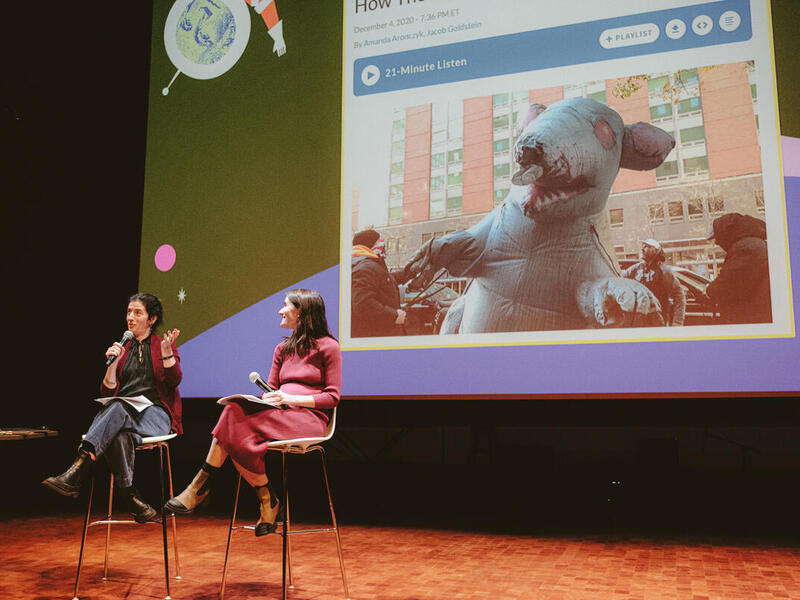 A phot of two women sitting in chairs on a stage in front of a large projection. The woman on the left is speaking into a microphone. The woman on the right is holding a microphone in her lap while watching the woman on the left speak.
