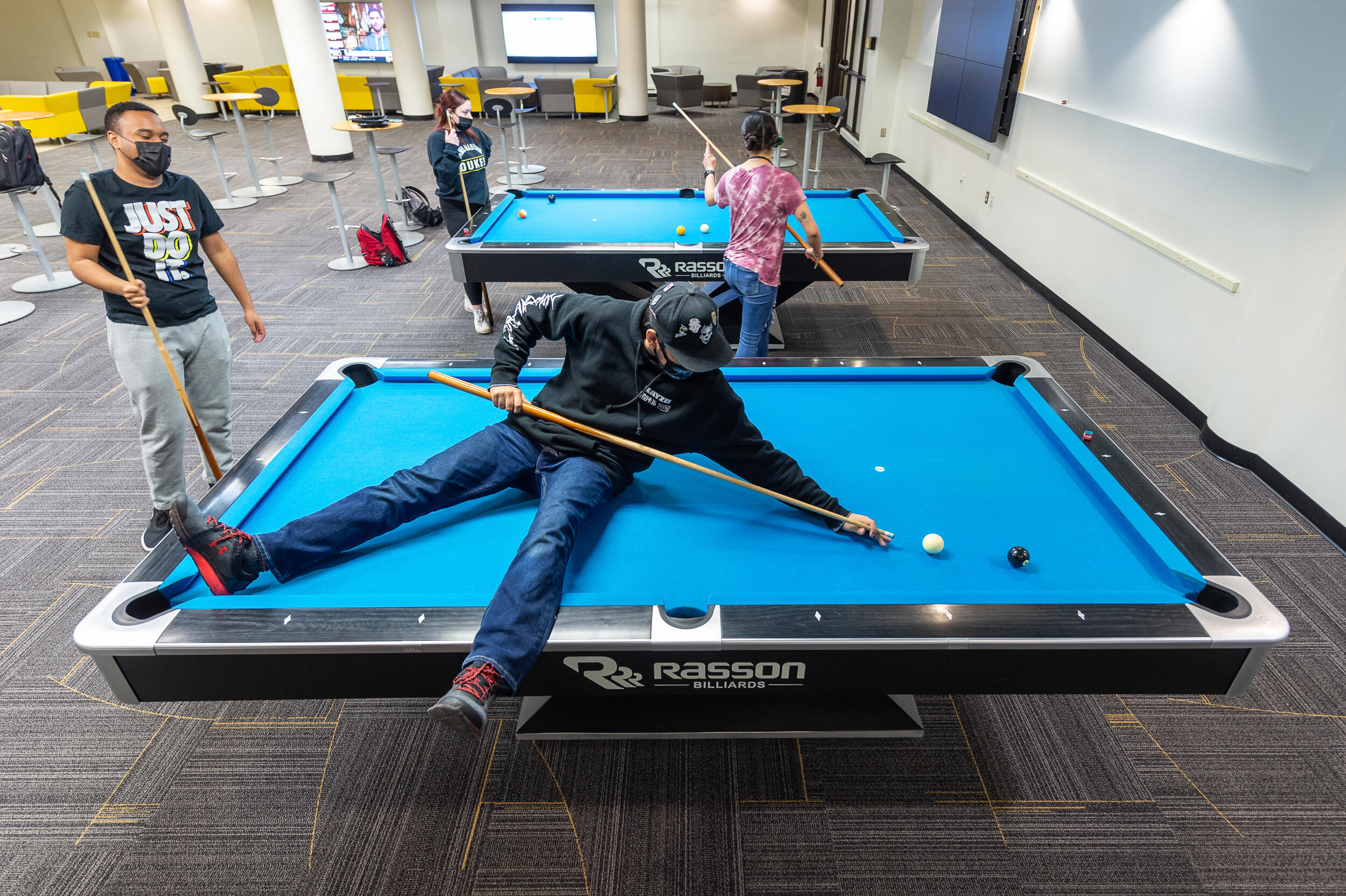 A student sits on a bright blue pool table in a large indoor space with other pool tables where several students wait to play pool.