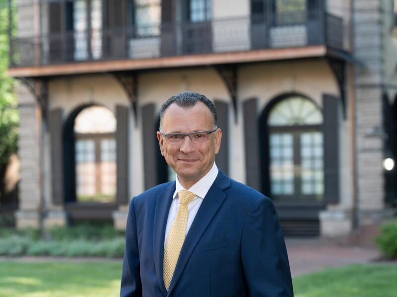 A man wearing a suit jacket, button down shirt, tie, and glasses while standing in front of a building. 