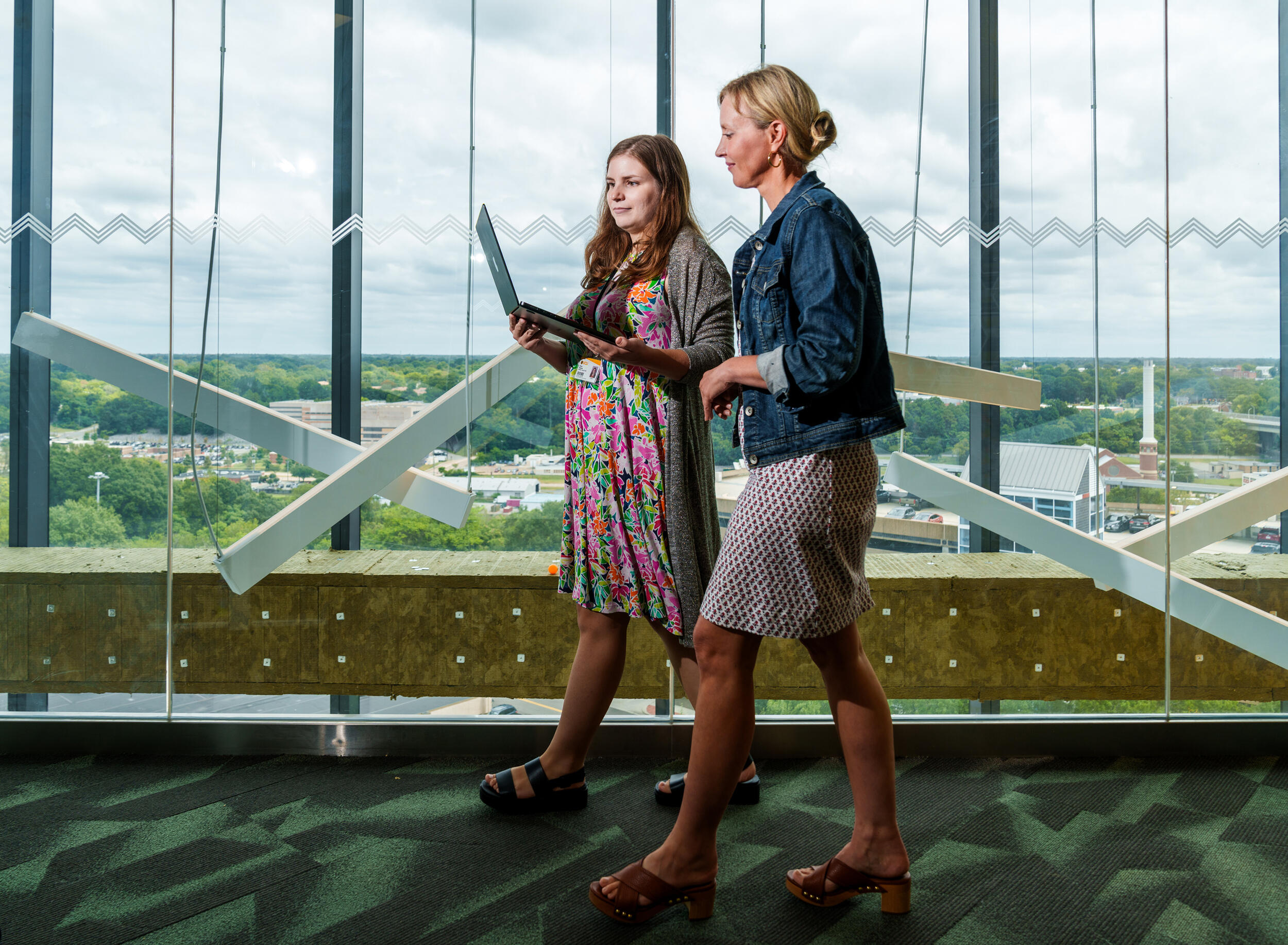 A photo of two women walking past a floor to ceiling window. The woman on the left is holding a laptop the woman on the right is looking at. 