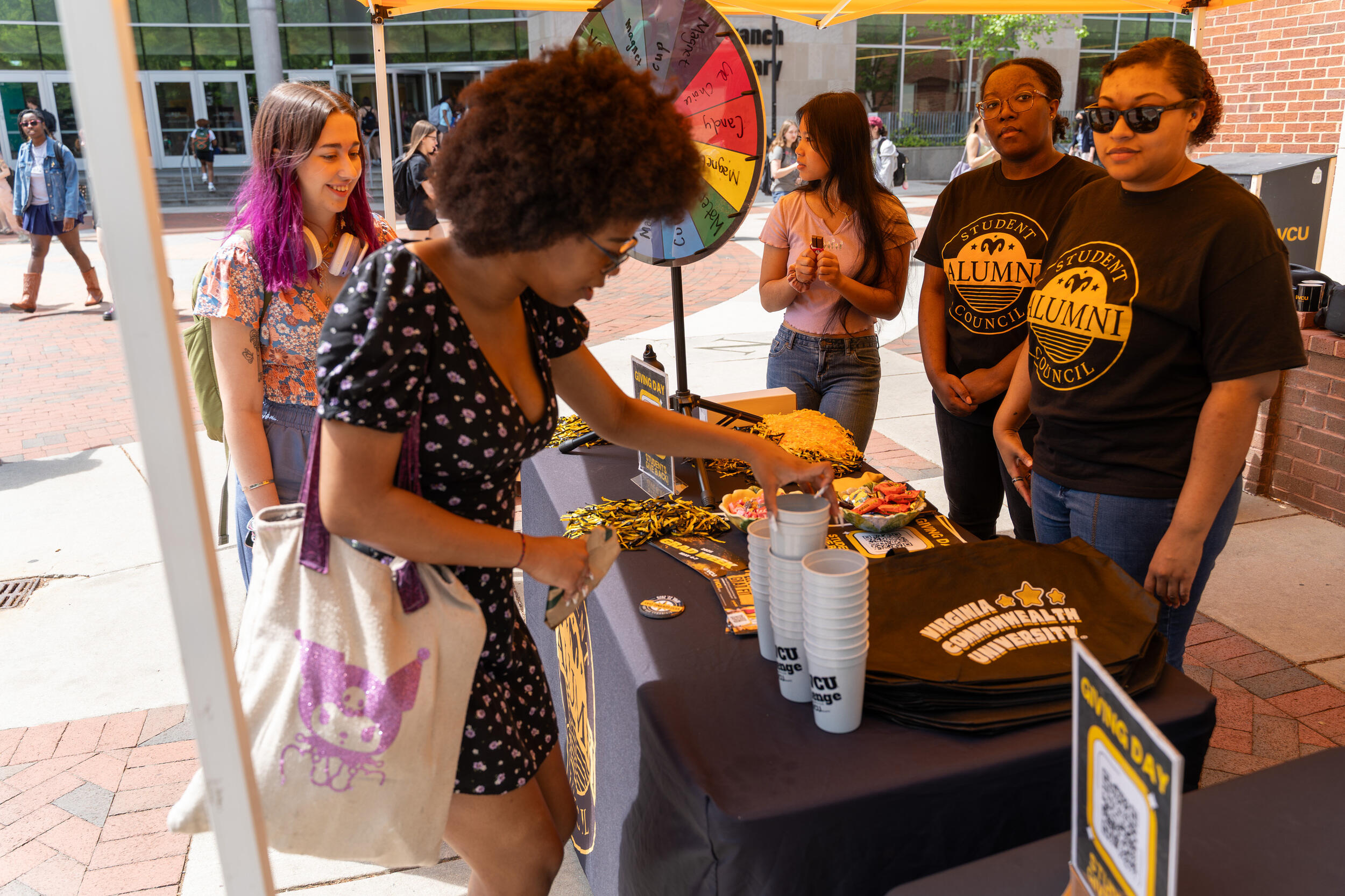 A photo of three women standing behind a table with and two women standing on the other side of it. The table has pom poms, cups, and other VCU-branded swag for people to take. To the left of the table is a multi colored prize wheel. 