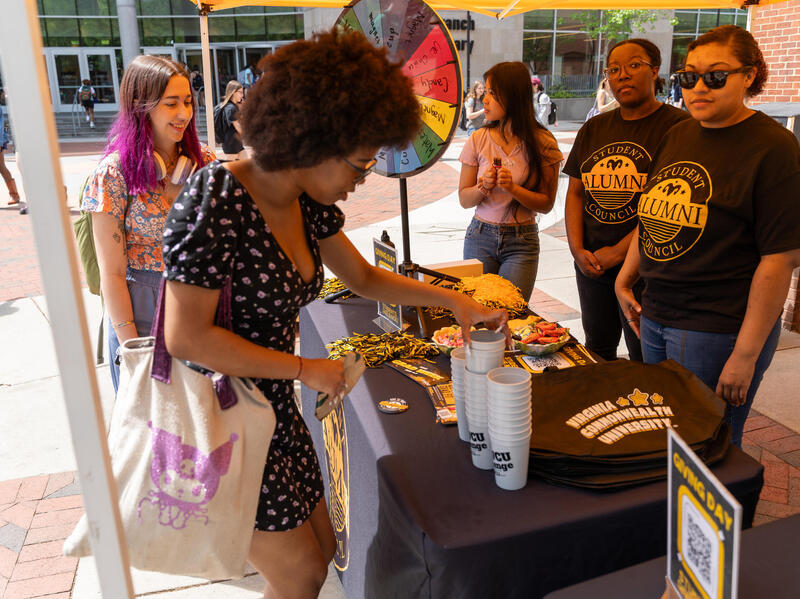 A photo of three women standing behind a table with and two women standing on the other side of it. The table has pom poms, cups, and other VCU-branded swag for people to take. To the left of the table is a multi colored prize wheel. 