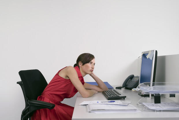 Woman in red dress sitting in a black office chair while slouched over a desk looking at a computer.