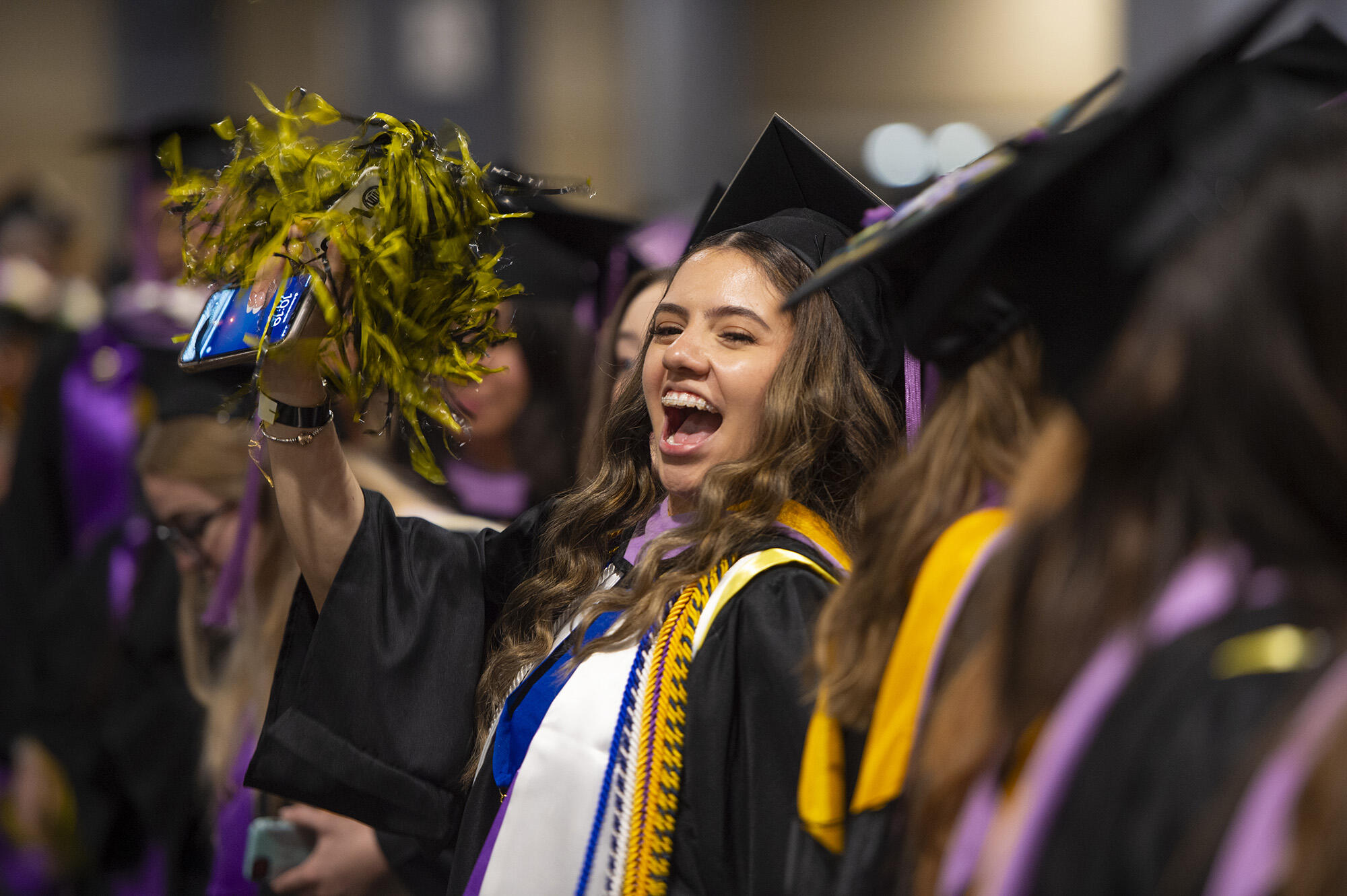 A photo of a woman wearing a graduation cap and gown holding a pompom and cheering.She is seated around many other graduating students who are blurry. 