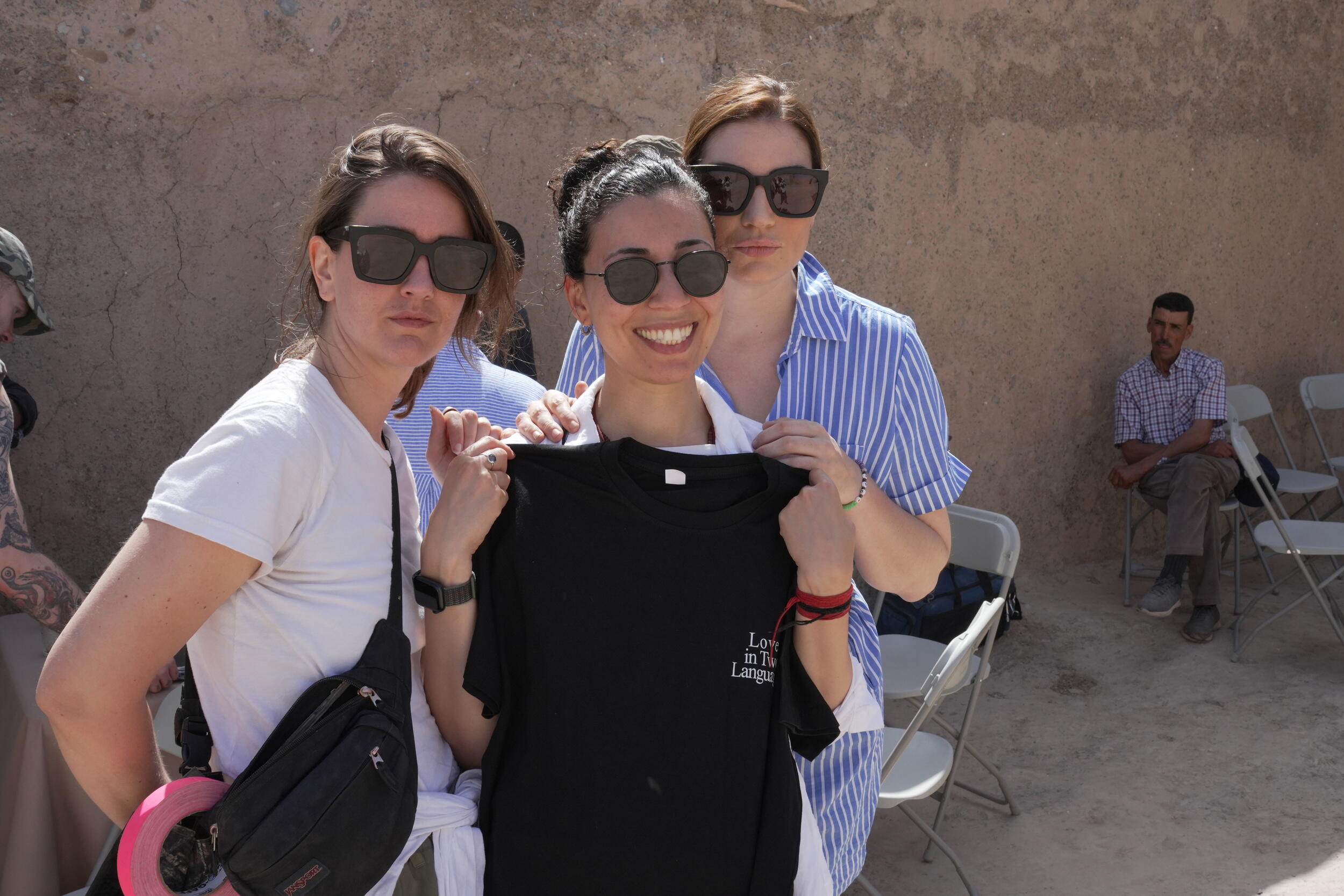 A photo of threde women standing outside. The woman in the center is holding a black t-shirt up to her chest. 