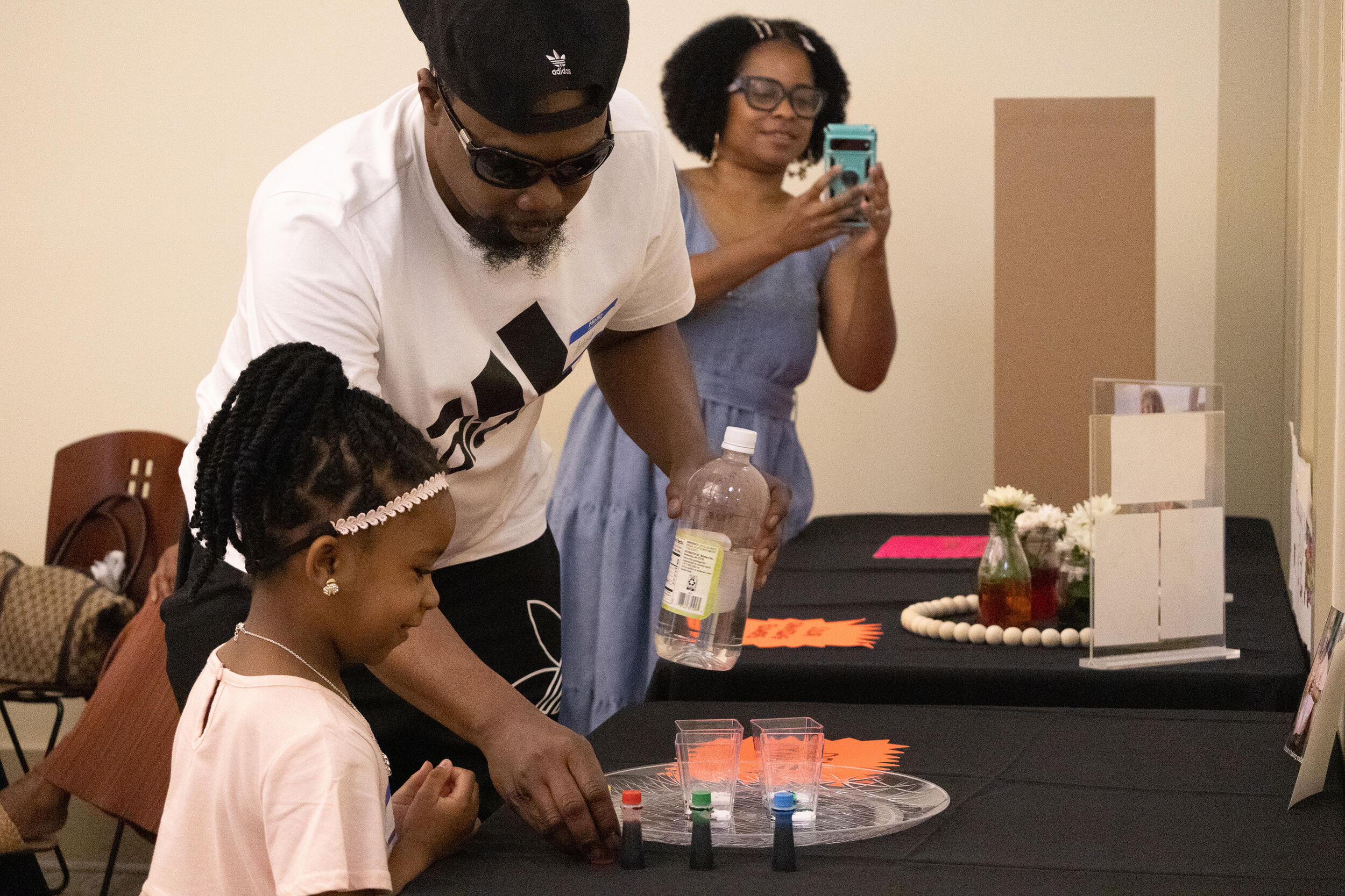 A photo of a man and a girl at a table looking at cups with different colored liquids in them. A woman is standing to the side holding her phone up. 