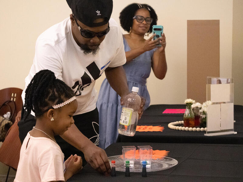 A photo of a man and a girl at a table looking at cups with different colored liquids in them. A woman is standing to the side holding her phone up. 