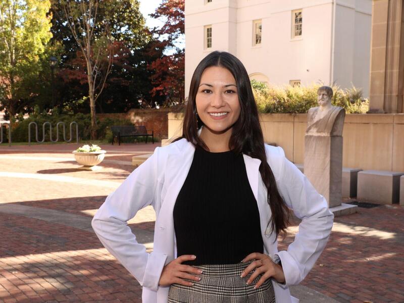 A photo of a woman standing outside with her hands on her hips. She is wearing a white lab coat and smiling. 