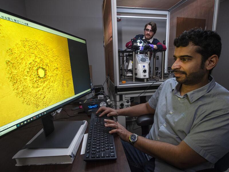 A photo of a man sitting in front of a computer monitor. A man standing to the right of him is looking at a machine. 