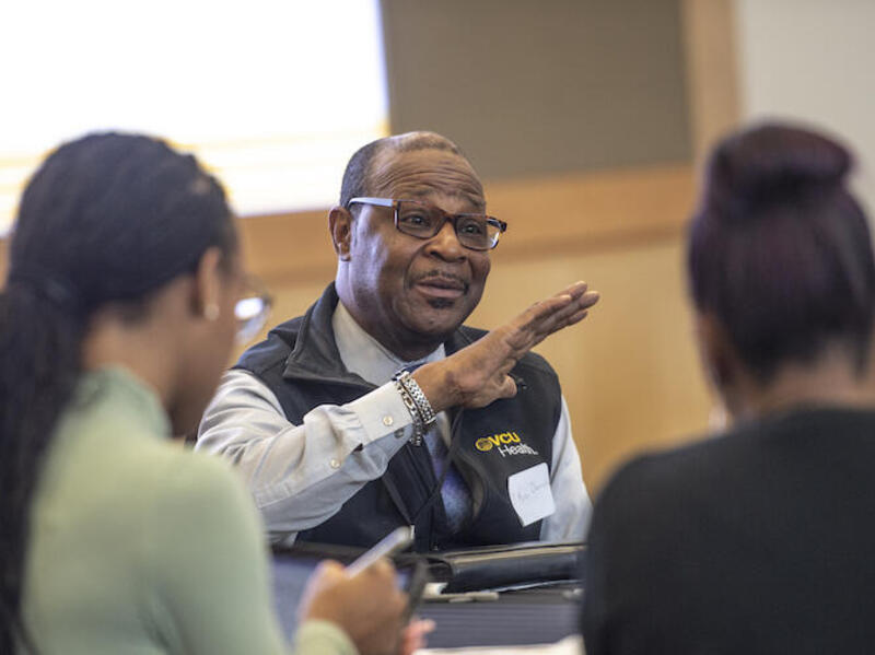 A photo of a man sitting at a table talking to two people sitting in front of him. 