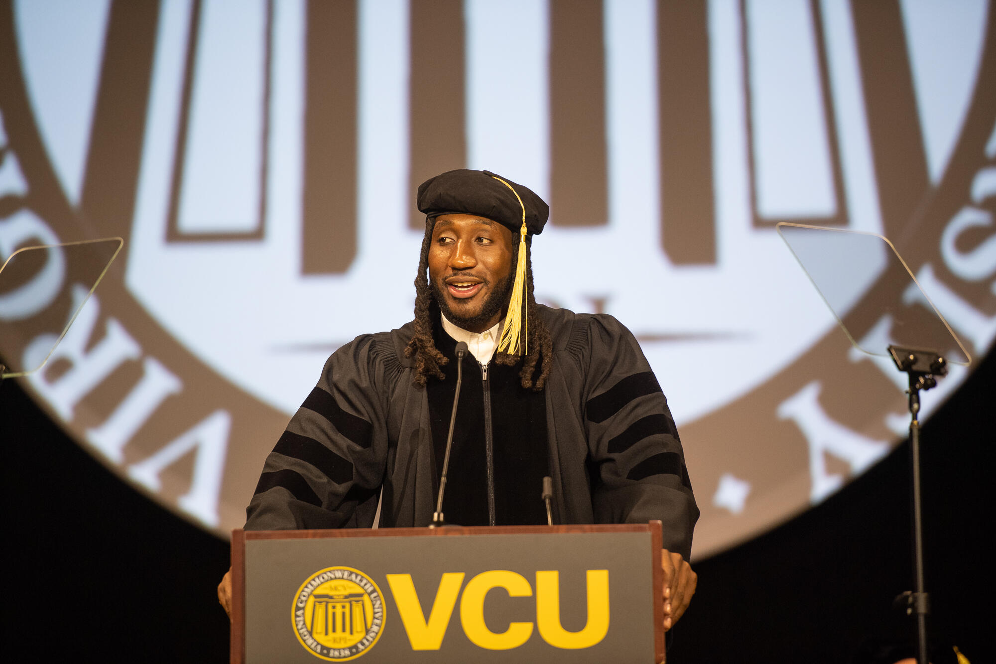 Mo Alie-Cox standing at the podium wearing commencement regalia.