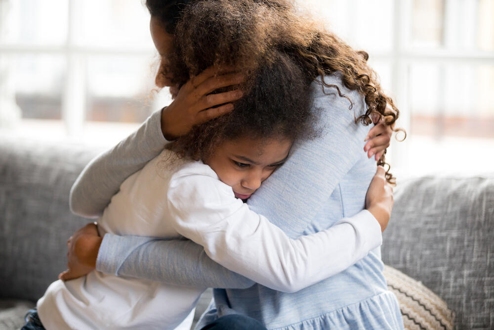 Mom in blue shirt comforts young, worried child with a hug. 