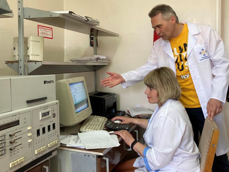 A woman is sitting at a desk on a computer while a man next to her has his arm outstretched pointing at it. 